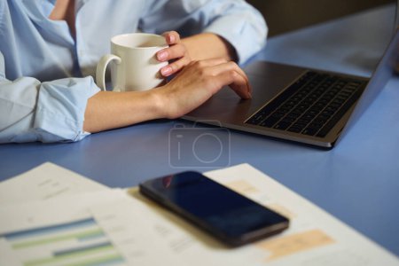 Photo for Cropped photo of woman with cup in hand seated at desk clicking on laptop touchpad - Royalty Free Image