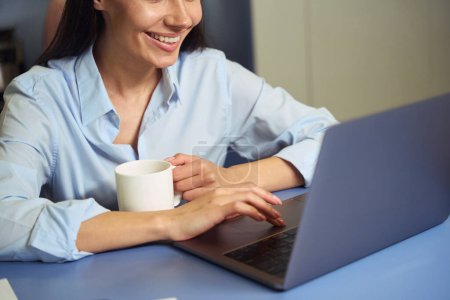 Téléchargez les photos : Photo recadrée d'une femme souriante avec une tasse à la main assise à la table en cliquant sur le pavé tactile de l'ordinateur portable - en image libre de droit