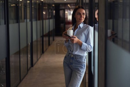 Photo for Dreamy company employee leaning against glass wall in hallway while holding ceramic teacup in hands - Royalty Free Image