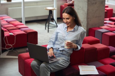 Photo for Pleased female freelancer with mug in hand looking at laptop screen while sitting on cozy chair in coworking space - Royalty Free Image