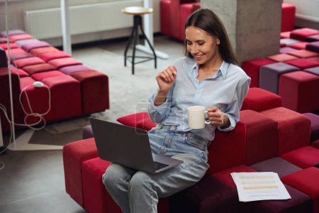 Téléchargez les photos : Femme souriante avec tasse à la main regardant quelque chose sur l'écran de l'ordinateur tout en étant assis sur une chaise dans un espace de coworking confortable - en image libre de droit