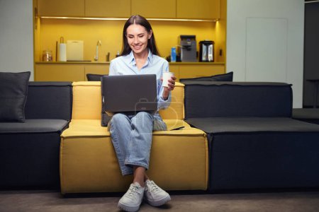 Téléchargez les photos : Travailleur d'entreprise souriant avec ordinateur portable et tasse en papier dans les mains assis sur le canapé dans la cuisine de bureau - en image libre de droit