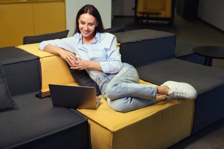 Téléchargez les photos : Jeune femme souriante regardant écran d'ordinateur portable tout en étant assis sur le canapé dans le salon avec tasse en papier à la main - en image libre de droit