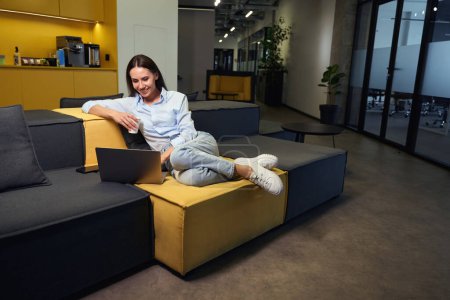 Téléchargez les photos : Joyeuse jeune femme avec ordinateur portable et tasse en papier jetable à la main assise sur le canapé dans la cuisine d'entreprise - en image libre de droit
