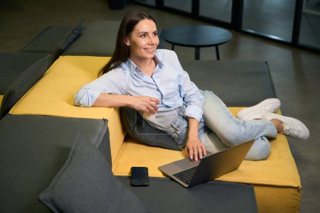 Téléchargez les photos : Jeune femme rêveuse avec tasse en papier à la main et ordinateur portable assis sur le canapé dans le salon - en image libre de droit