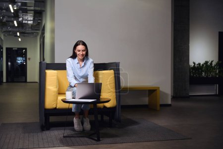 Photo for Smiling office worker typing on computer keyboard while sitting at coffee table in lounge area - Royalty Free Image
