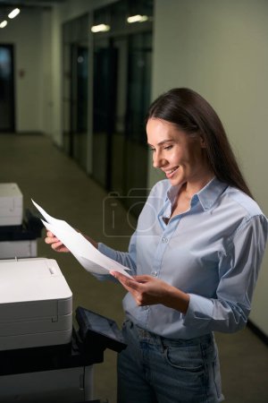 Foto de Agradable empleada corporativa femenina mirando fotocopias en sus manos mientras está de pie frente a la fotocopiadora en el pasillo - Imagen libre de derechos