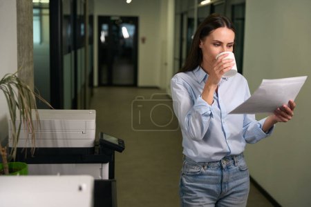 Foto de Trabajadora de oficina enfocada examinando fotocopias mientras está de pie junto a la fotocopiadora en el pasillo - Imagen libre de derechos
