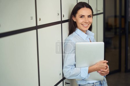 Téléchargez les photos : Portrait taille haute d'un employé joyeux de l'entreprise avec un ordinateur portable appuyé contre une armoire de rangement - en image libre de droit