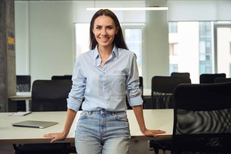 Photo for Joyous young company employee leaning against desk in open-plan office and looking in front of her - Royalty Free Image
