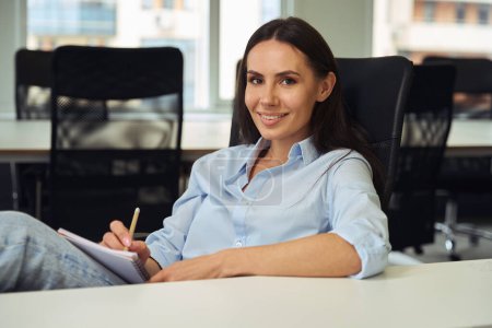 Téléchargez les photos : Travailleur d'entreprise souriant avec crayon et journal dans les mains assis au bureau dans un bureau décloisonné - en image libre de droit