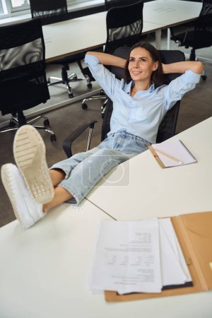 Foto de Sonriente empleada corporativa soñadora sentada en una oficina de planta abierta con los pies sobre la mesa - Imagen libre de derechos