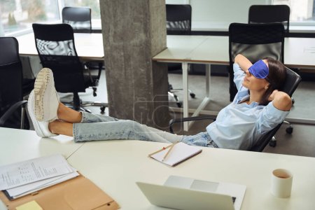 Téléchargez les photos : Calme employée d'entreprise avec masque de sommeil sur les yeux assis dans un bureau décloisonné avec ses pieds sur la table - en image libre de droit