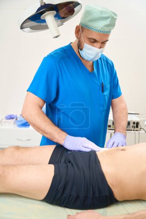 Foto de Cirujano masculino que aplica un yeso adhesivo en el estómago de los pacientes después de las inyecciones. Tratamiento venoso de malla - Imagen libre de derechos