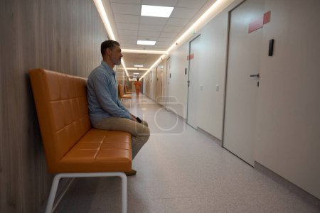 Photo for Adult male is waiting on a brown couch outside a doctor office in the hospital long corridor. - Royalty Free Image