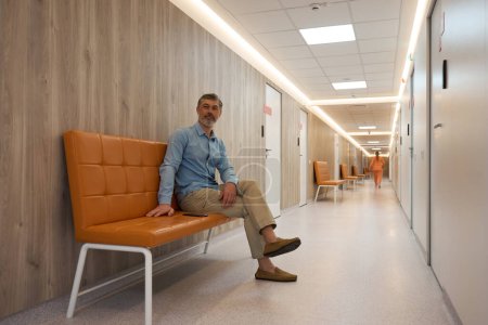 Photo for Gray-haired adult man sitting on a brown sofa in the corridor of a medical center with wooden walls - Royalty Free Image