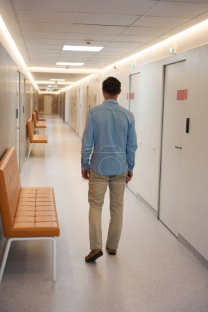 Téléchargez les photos : Homme debout dans le couloir de la clinique médicale avec des canapés bruns le long du mur. Vue arrière - en image libre de droit