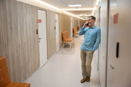 Photo for Adult man with beard leaning against the wall is talking on the phone in hallway of the clinic. Full height - Royalty Free Image