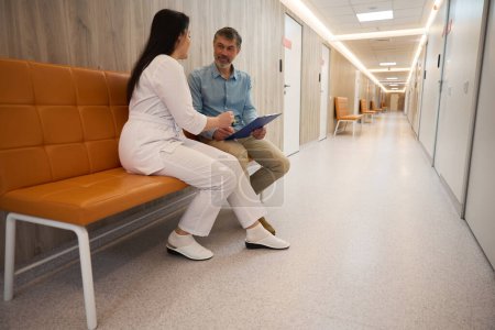 Foto de Mujer trabajadora médica en traje blanco se sienta con el paciente en el sofá y ayuda a llenar el cuestionario en el hospital - Imagen libre de derechos