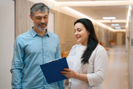 Photo for Smiling medical worker showing a folder with a questionnaire to a patient while standing in a well-lit corridor of the clinic - Royalty Free Image