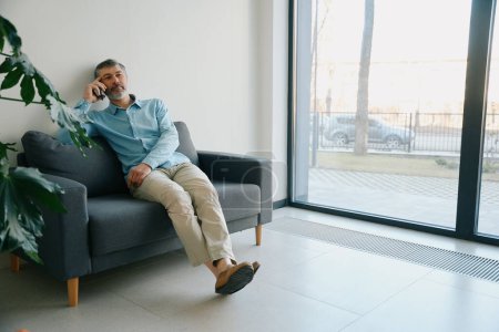 Téléchargez les photos : Homme assis sur un canapé noir près d'une grande fenêtre et des plantes d'intérieur dans le couloir de la clinique - en image libre de droit