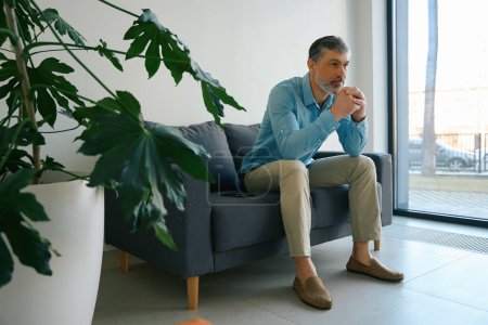 Téléchargez les photos : Homme aux cheveux gris avec une barbe appuyée sur ses mains s'assoit sur un canapé noir près de la fenêtre - en image libre de droit