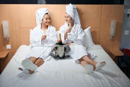 Foto de Bonitas señoritas multirraciales con una toalla en la cabeza y con abrigos blancos se sientan en la cama en el hotel y sostienen copas de champán - Imagen libre de derechos