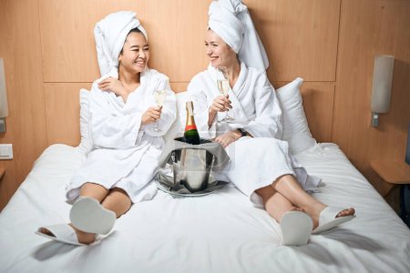 Foto de Dos mujeres en albornoces beben champán en una habitación de hotel. Celebra el viaje en la cama del hotel - Imagen libre de derechos