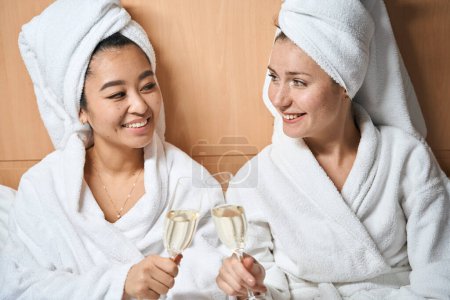 Foto de Dos novias con albornoces en la cama bebiendo champán. Una pequeña fiesta en una habitación de hotel - Imagen libre de derechos