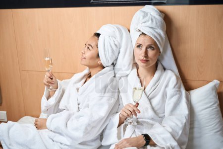 Foto de Mujeres en albornoces y con champán en la cama. Dos amigas vestidas de blanco con gafas en un hotel - Imagen libre de derechos