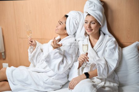 Foto de Novias en albornoces y con champán en el hotel. Dos mujeres en abrigos blancos con gafas en la cama - Imagen libre de derechos