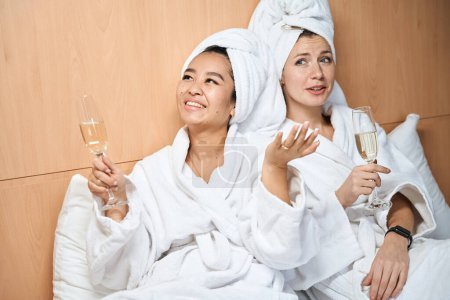 Foto de Felices amigas bebiendo champán con batas blancas. Dos damas en el hotel bebiendo champán en la cama - Imagen libre de derechos