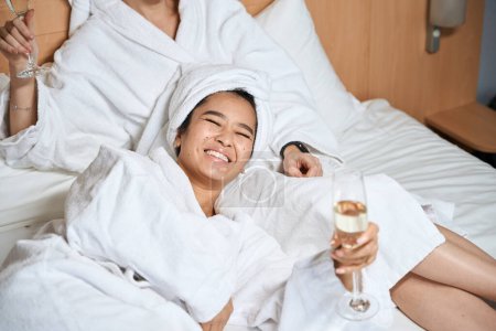 Téléchargez les photos : Joyeuses dames buvant du champagne dans la chambre d'hôtel. Deux amies en peignoirs blancs boivent du champagne sur le lit - en image libre de droit