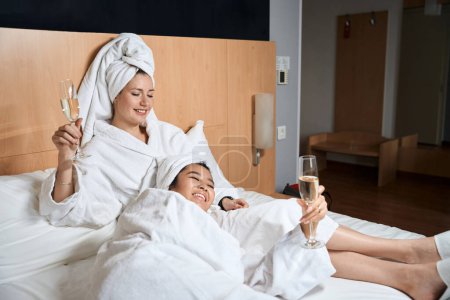 Téléchargez les photos : Deux dames heureuses boivent du champagne en peignoirs blancs au lit. Copines avec des lunettes dans les mains dans une chambre d'hôtel - en image libre de droit