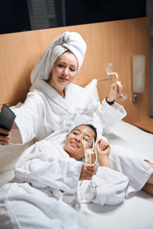 Téléchargez les photos : Deux dames allongées sur le lit prennent selfie dans la chambre d'hôtel. Les amies se reposent sur le lit dans la chambre d'hôtel - en image libre de droit