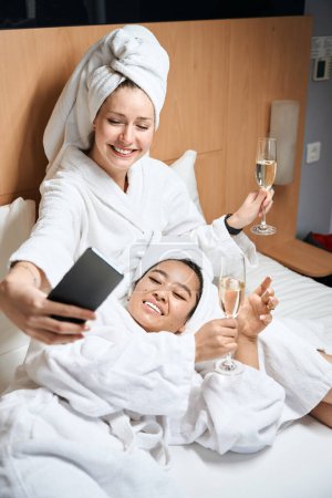 Photo for Two girlfriends are photographed on the phone in bathrobes. Ladies in white coats with glasses take a selfie - Royalty Free Image