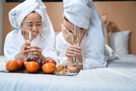 Téléchargez les photos : Deux copines boivent du champagne avec des fruits en manteaux blancs. Les jeunes femmes après une douche célèbrent dans une chambre d'hôtel - en image libre de droit