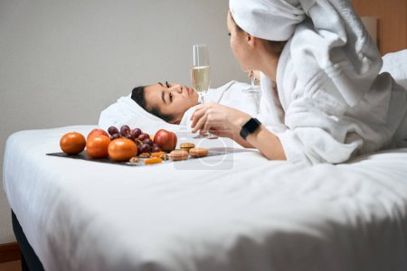 Foto de Vista lateral de una hembra con toallas en la cabeza se encuentran en la cama con frutas y champán. despedida de soltera en el hotel - Imagen libre de derechos