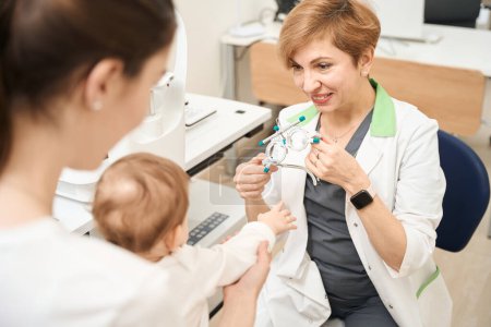 Téléchargez les photos : Un ophtalmologiste souriant tenant un cadre d'essai devant un bébé et un parent assis dans son bureau - en image libre de droit