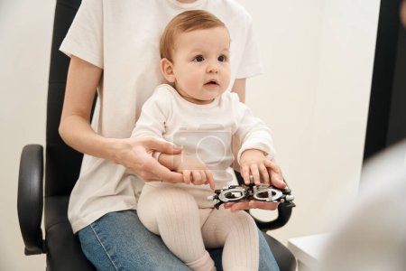 Photo for Cropped photo of woman with ophthalmic trial frame in hand and calm little child on lap sitting at optometrist office - Royalty Free Image