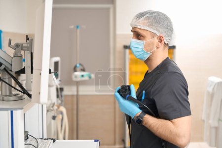 Photo for Side view of focused endoscopist with endoscope in gloved hand looking at monitor screen - Royalty Free Image
