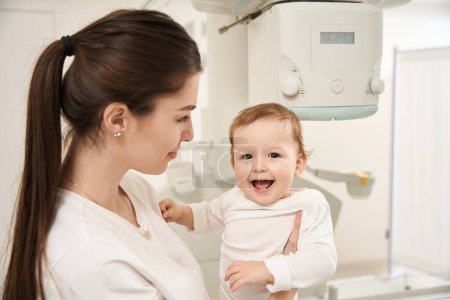 Foto de Joven madre sosteniendo a su pequeño bebé complacido frente a un aparato de rayos X - Imagen libre de derechos