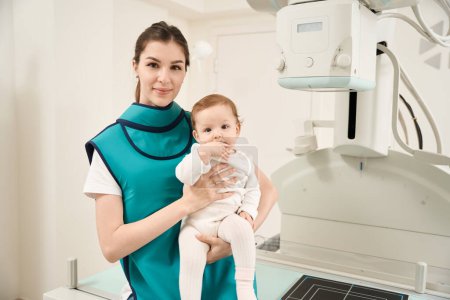 Photo for Smiling woman in lead apron holding calm baby in her arms in front of x-ray machine - Royalty Free Image
