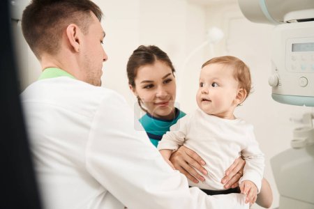 Foto de Técnico de rayos X que coloca un escudo pélvico en un niño pequeño en presencia de padres femeninos satisfechos - Imagen libre de derechos