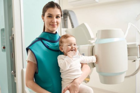 Téléchargez les photos : Agréable jeune maman tenant gai petit enfant dans ses bras tout en se tenant à côté de la machine à rayons X - en image libre de droit
