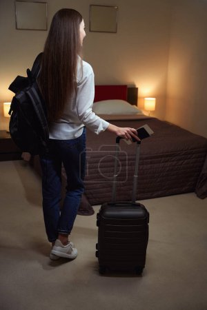 Foto de Señora sosteniendo una maleta por el mango mientras está de pie en una habitación de hotel. Vista desde atrás - Imagen libre de derechos