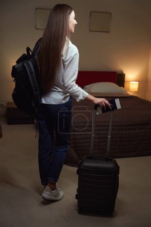 Téléchargez les photos : Femme tenant un bagage par la poignée alors qu'elle se tenait dans une chambre d'hôtel. Vue de l'arrière - en image libre de droit