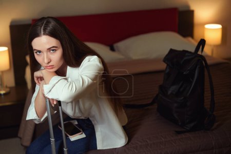 Téléchargez les photos : Portrait d'une jeune femme triste assise sur son lit appuyée sur une valise dans une chambre d'hôtel - en image libre de droit