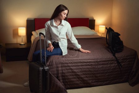 Téléchargez les photos : Jolie jeune femme assise sur le lit et redressant la couverture d'une main dans la chambre d'hôtel. Check-in hôtel - en image libre de droit
