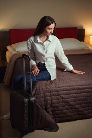 Téléchargez les photos : Femme lissant le couvre-lit assis sur un grand lit avec une valise à côté d'elle dans une chambre d'hôtel - en image libre de droit